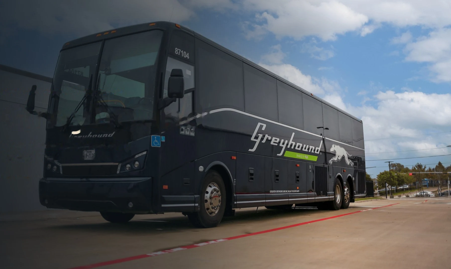 A dark Greyhound coach parked on a road under a blue sky with clouds.